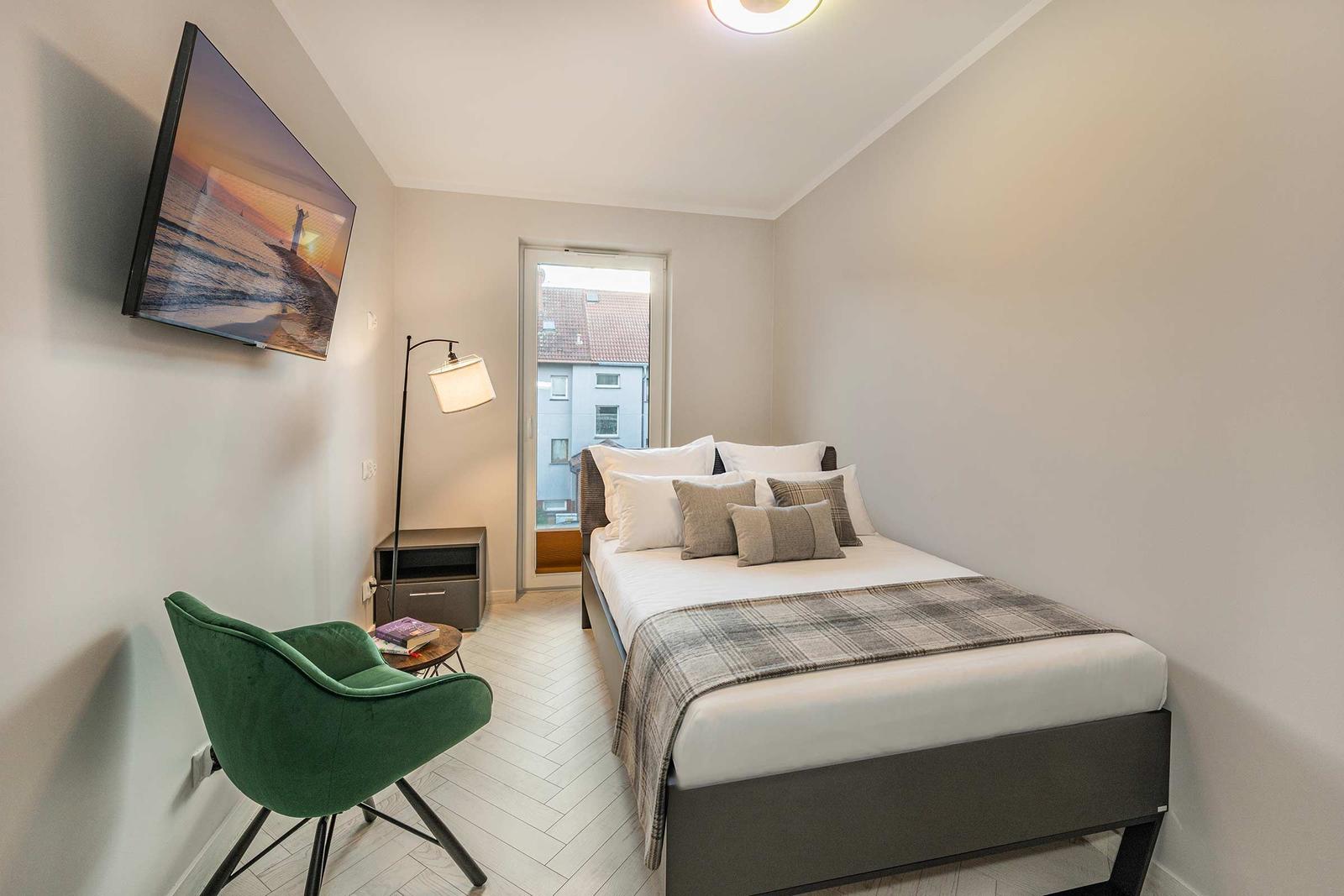 Schlafzimmer mit Bett, grüner Sessel und Wandbild. Tür führt nach draußen.