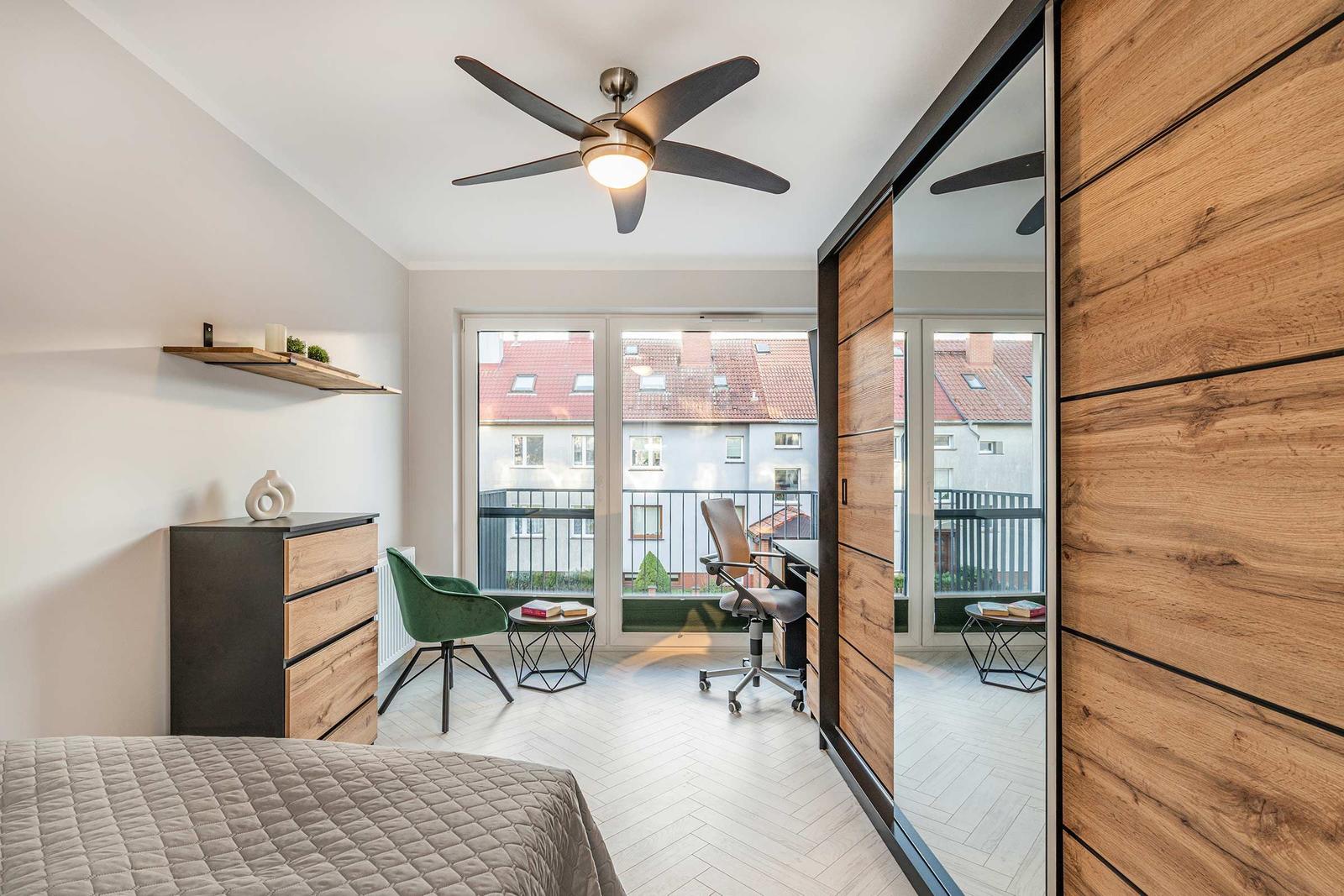Schlafzimmer mit Balkon, Schreibtisch und Holzschrank. Deckenventilator und Wandregal sichtbar.