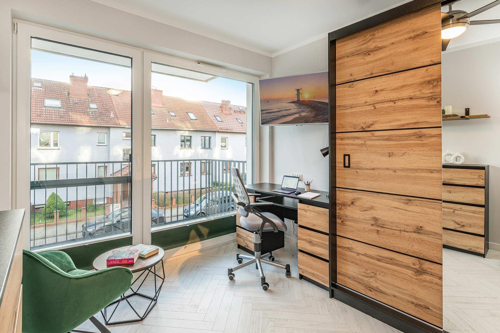 Zimmer mit Schreibtisch, grüner Sessel und Balkon mit Blick auf Wohnhäuser.