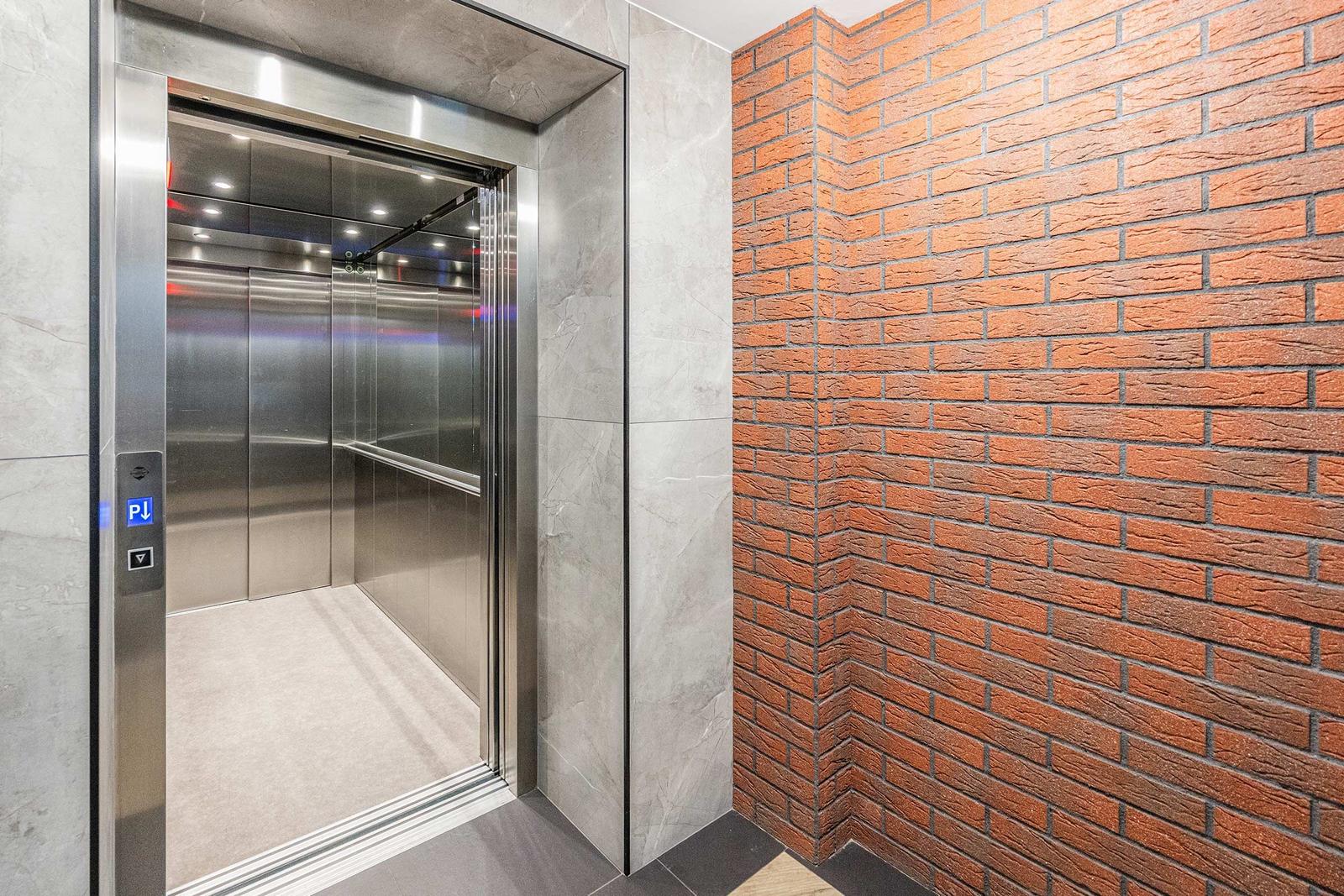 Elevator with stainless steel interior and control panel. Brick wall and marble tiles surround the entrance.