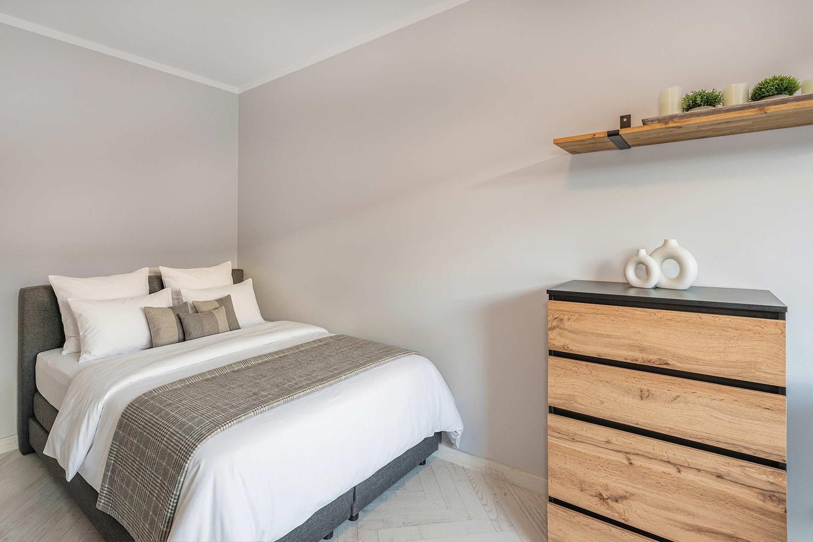 Schlafzimmer mit Bett, Schrank und Regal. Wandfarbe hellgrau. Boden hell gefliest.