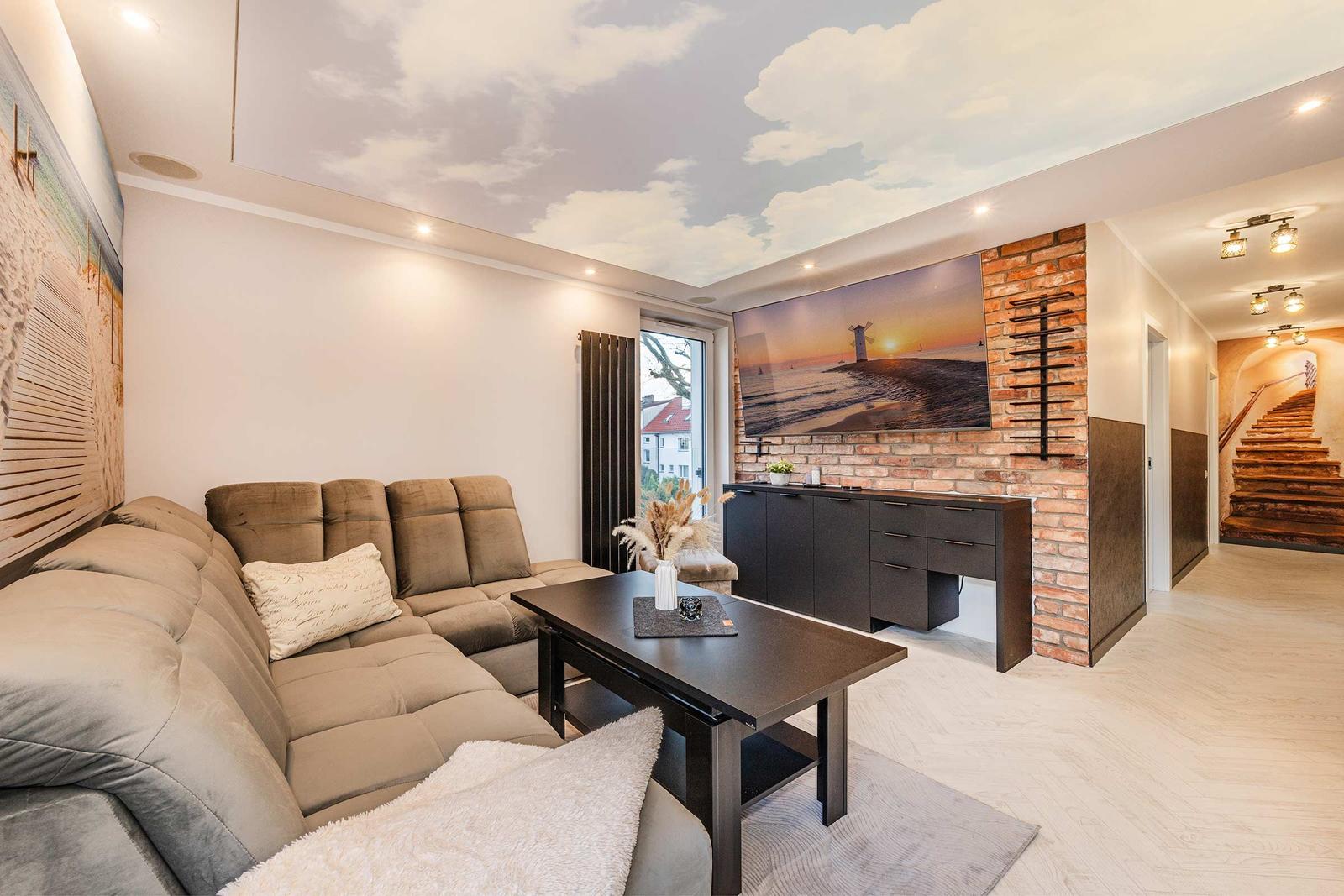 Wohnzimmer mit Sofa, Couchtisch und Wandbild. Decke mit Wolkenmotiv. Holzboden und Treppenhaus im Hintergrund.