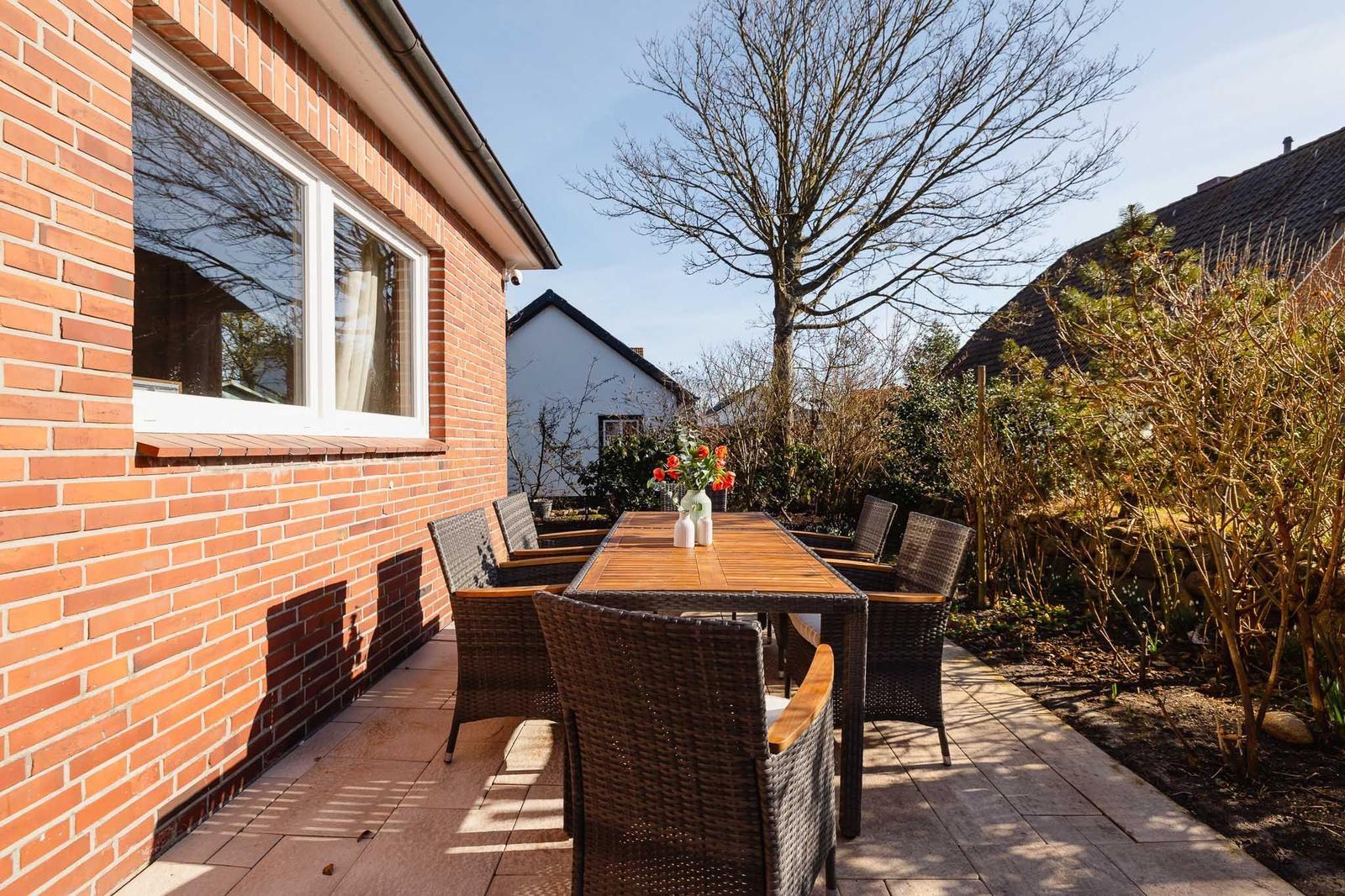 Terrasse mit Holztisch und Stühlen, roter Backsteinmauer und Gartenbereich.
