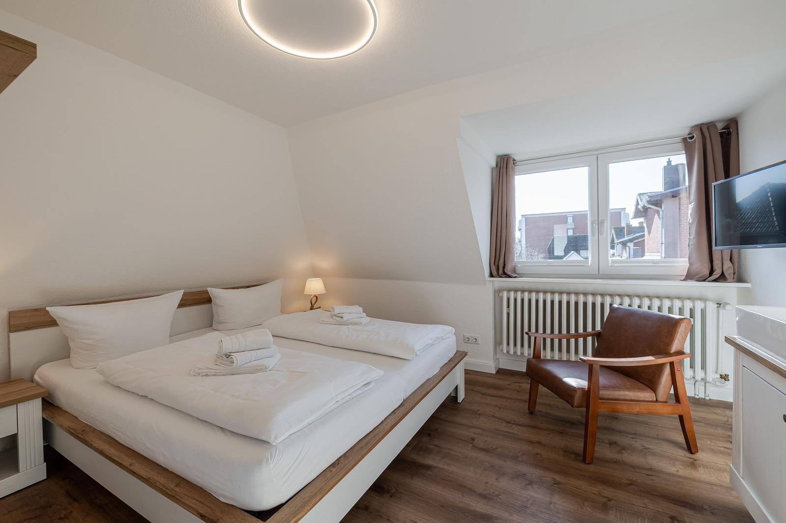Schlafzimmer mit Doppelbett, Sessel und Fenster. Holzboden und weiße Wände.