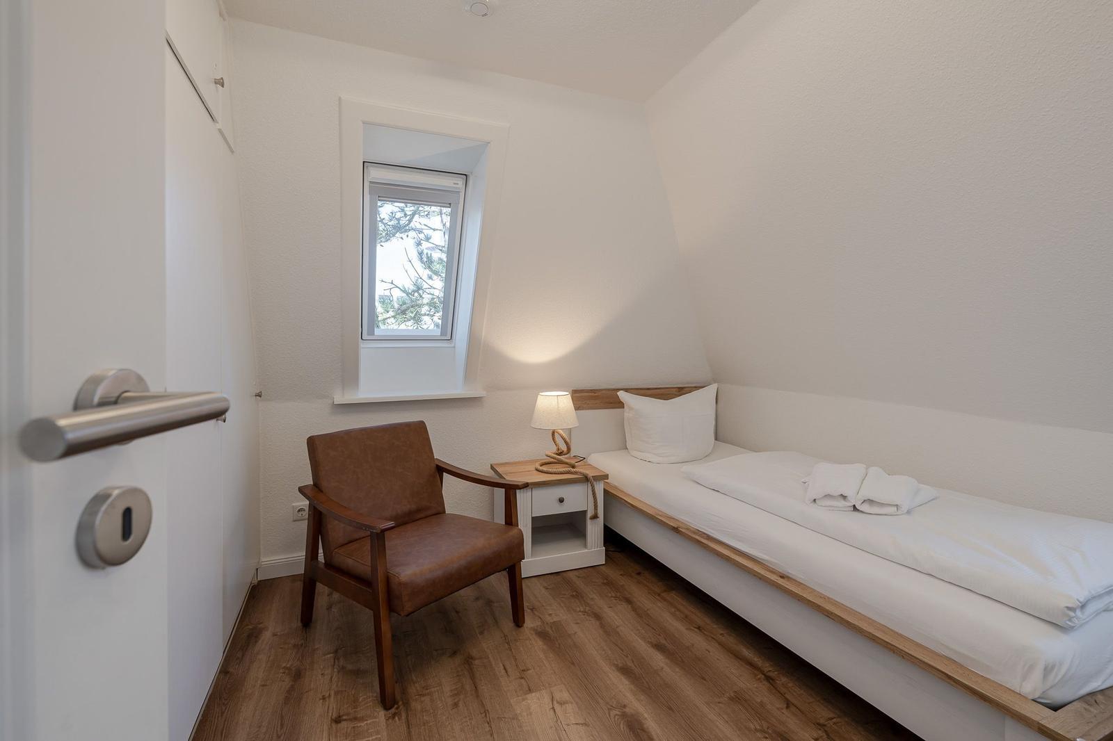 Schlafzimmer mit Bett, Stuhl und Fenster. Holzfußboden und weiße Wände.