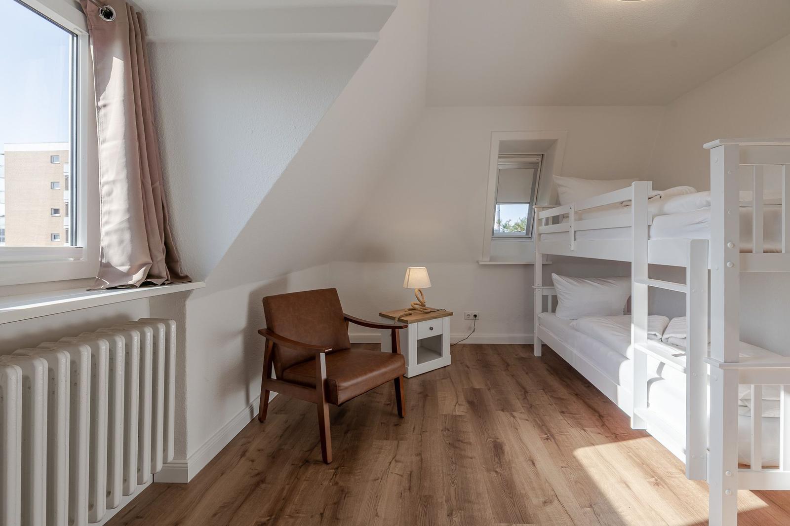 Schlafzimmer mit Doppelstockbett, brauner Sessel und Holzboden. Fenster mit Vorhang und Heizkörper.