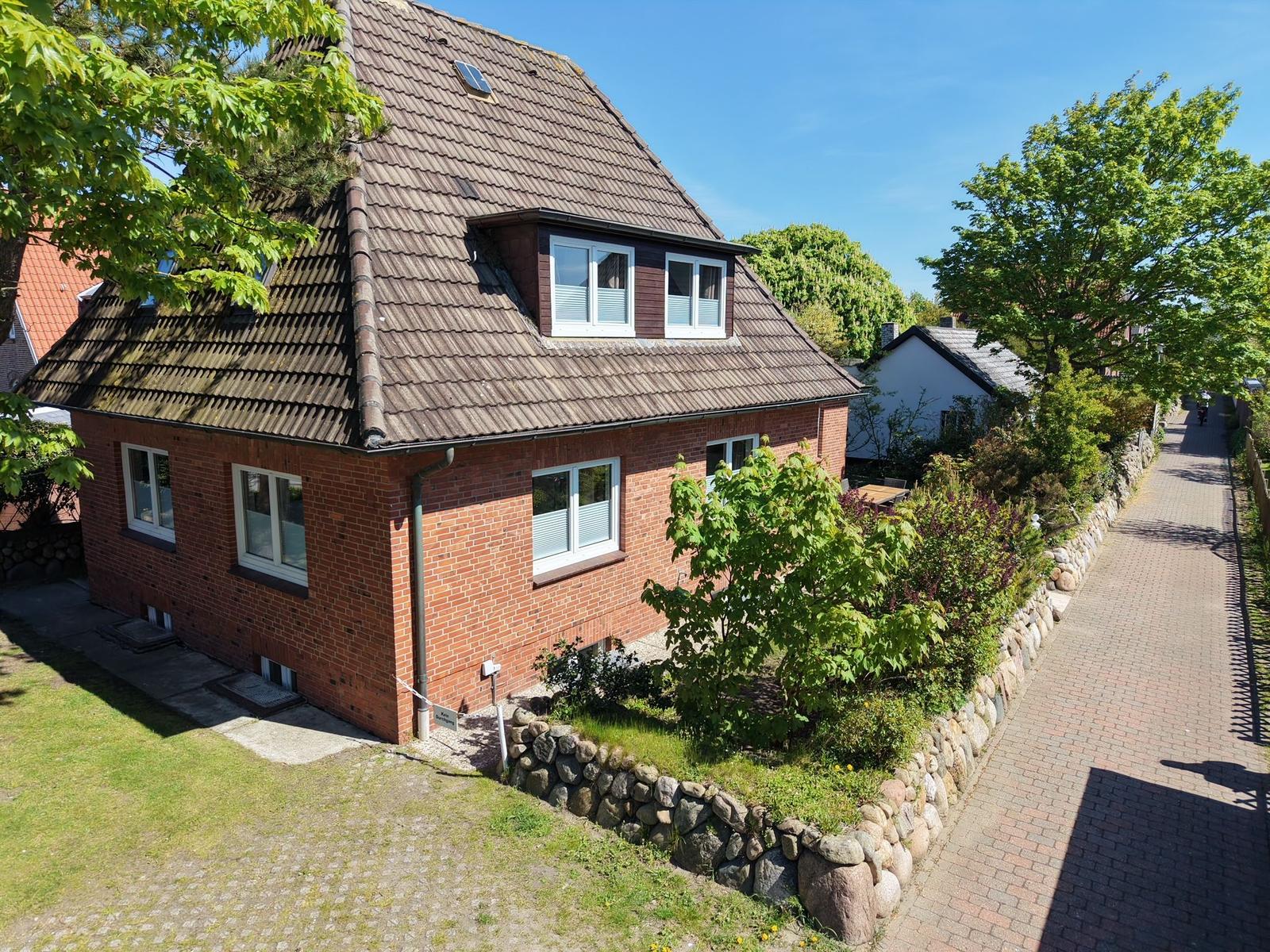 Rotes Backsteingebäude mit Dachgaube und Garten. Pflasterweg und Bäume umgeben die Immobilie.