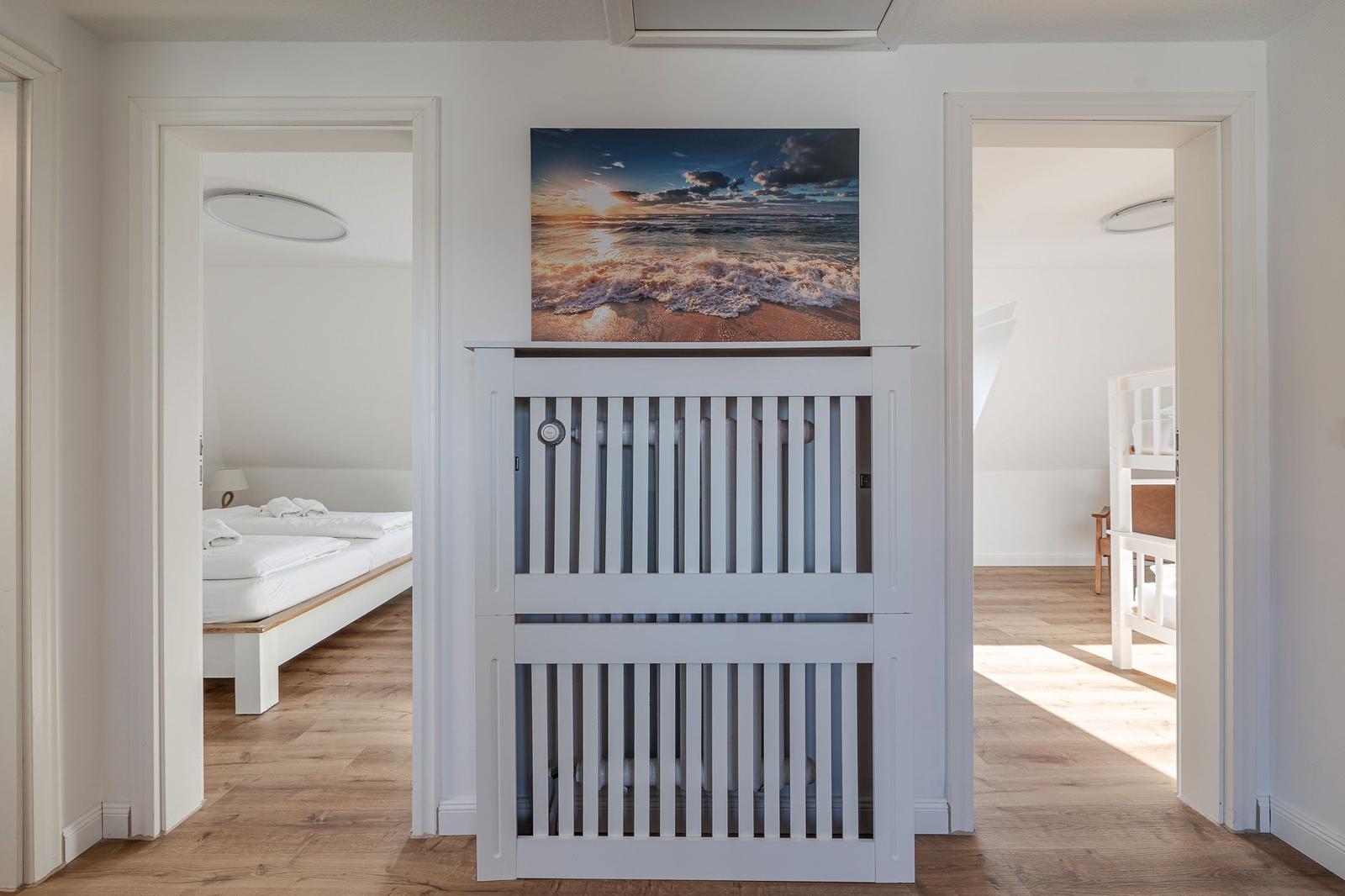 Hinter einer weißen Trennwand sind Schlafzimmer mit Bett und Kinderbett sichtbar. Über dem Tor hängt ein Strandbild.