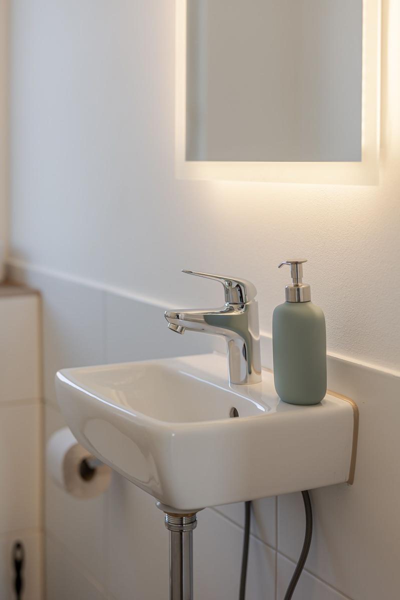 Waschbecken mit Armatur und Seifenspender unter beleuchtetem Spiegel.