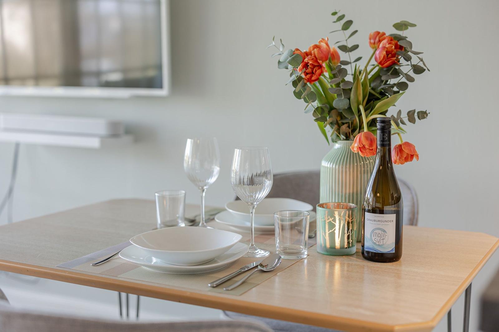 Gedeckter Esstisch mit Blumen, Wein und Geschirr. Hintergrund: Fernseher und Wandheizung.