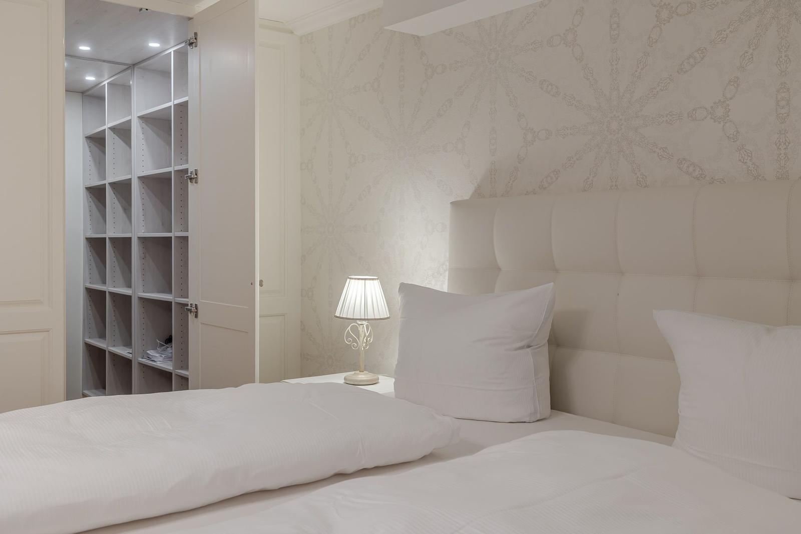 Schlafzimmer mit weißem Bett, Kopfteil und Nachttisch. An der Wand hängt ein Schrank mit offenen Fächern.