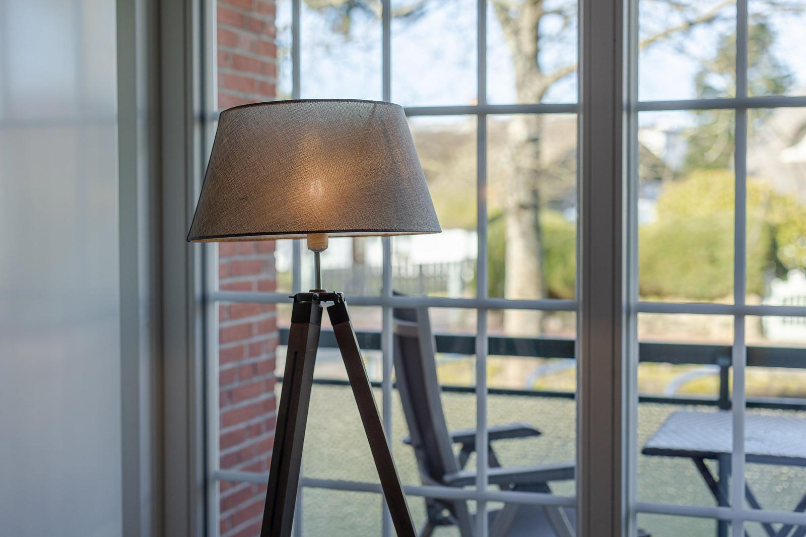 Eine Stehlampe mit Holzgestell steht vor einem Fenster mit Blick auf eine Terrasse.