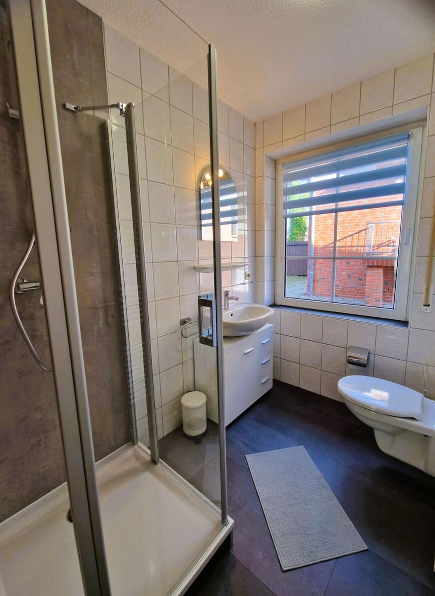 Badezimmer mit Dusche, Waschbecken und Toilette. Fenster mit Jalousie.