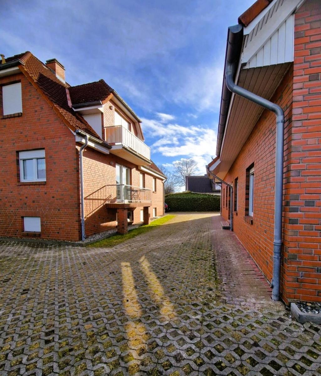 Zweistöckiges Haus mit Balkon und gepflastertem Hof. Hinterhof mit Hecke und blauem Himmel.