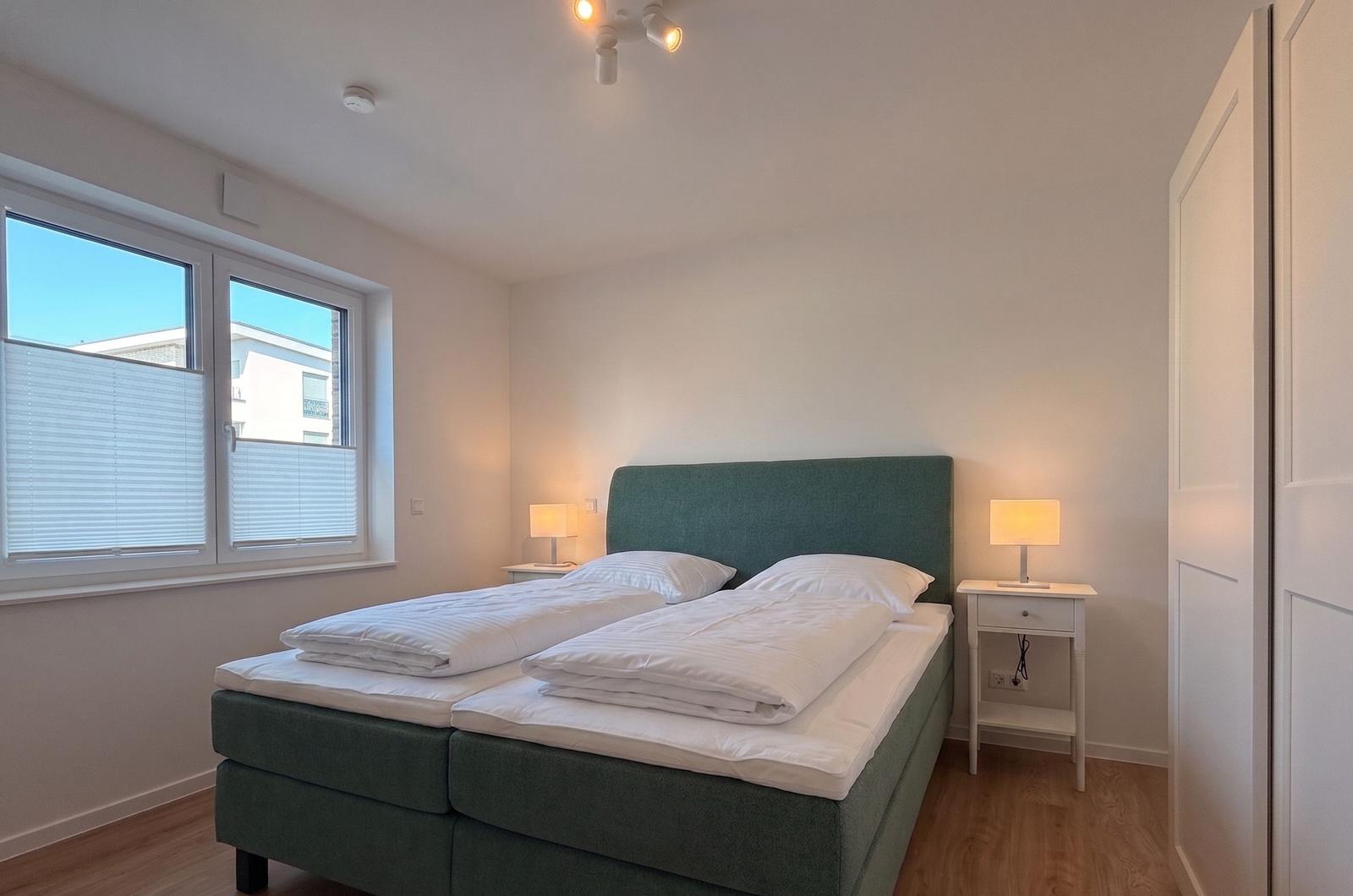 Schlafzimmer mit Bett, Nachttischen und Fenster. Holzboden und weiße Wände.