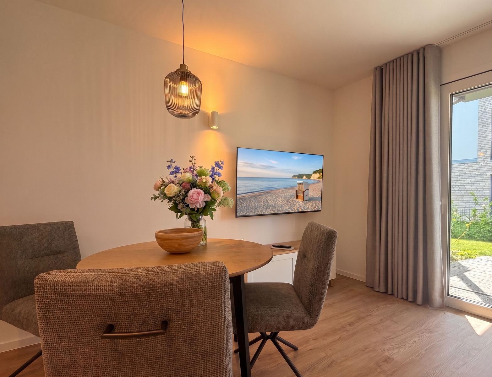 Rundes Esstisch mit Stühlen, Fernseher und Blumen auf Tisch. Terrasse mit Blick nach draußen.