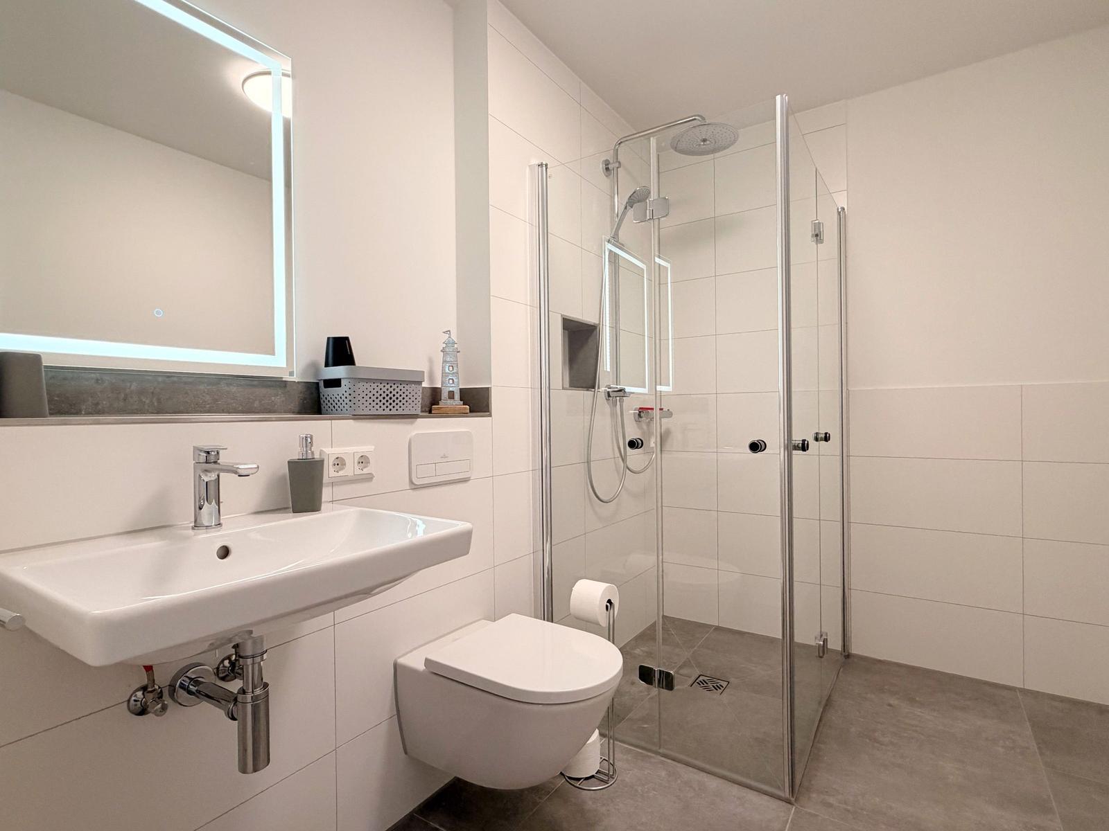 Badezimmer mit Dusche, Waschbecken und WC. Fliesenwand und Spiegel mit Beleuchtung.