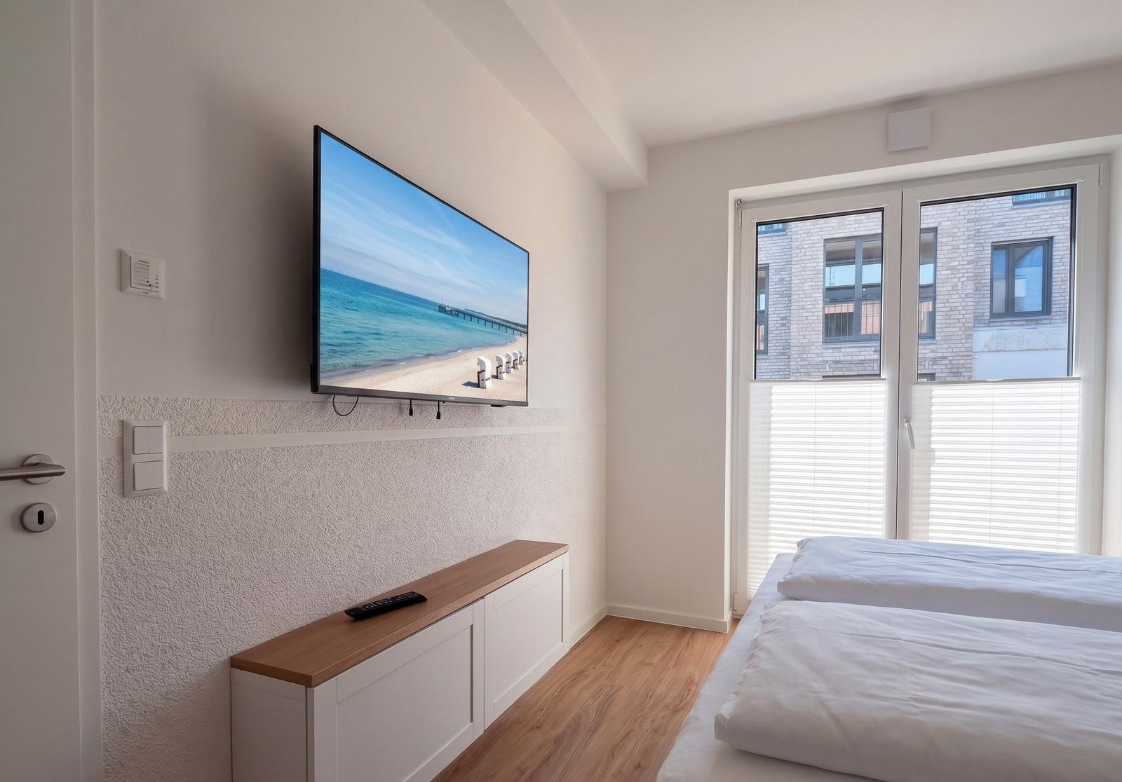 Schlafzimmer mit Bett, TV und Fenster. TV zeigt Strandbild.