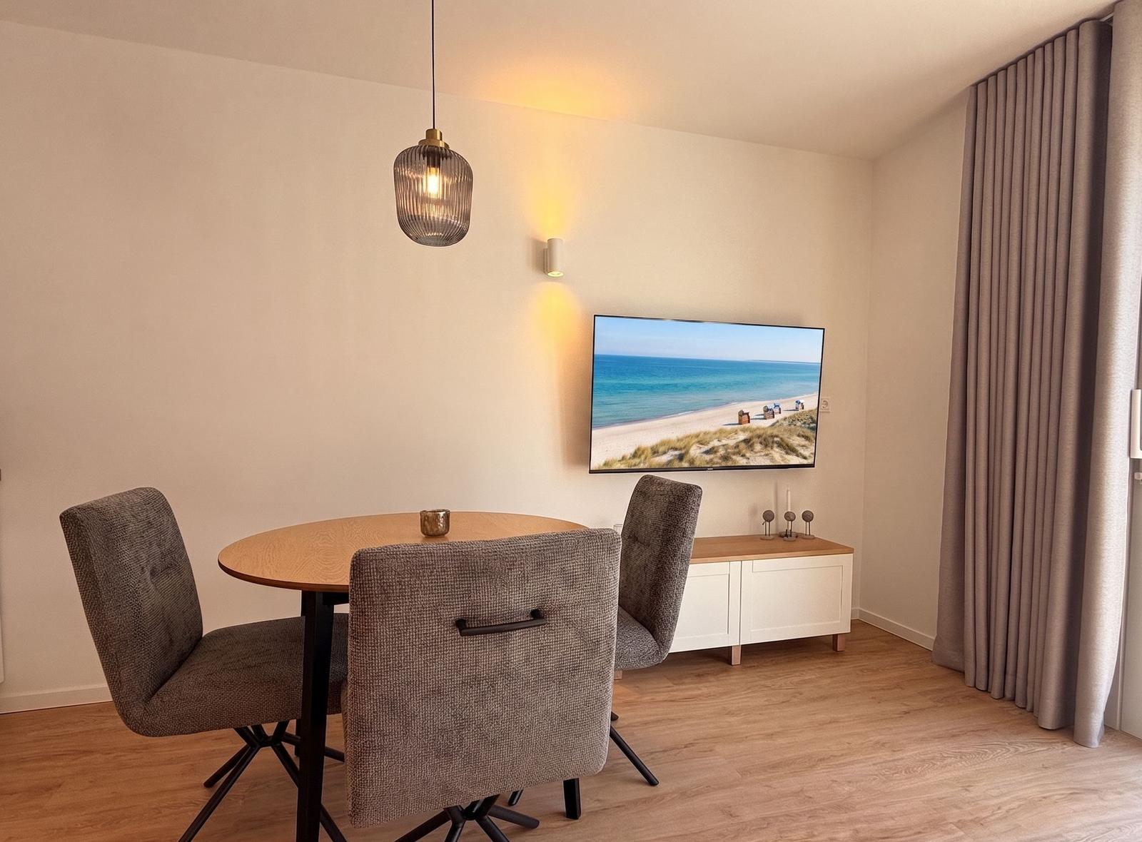 Essbereich mit runden Tisch und drei Stühlen. Fernseher zeigt Strand. Holzfußboden und Vorhänge.