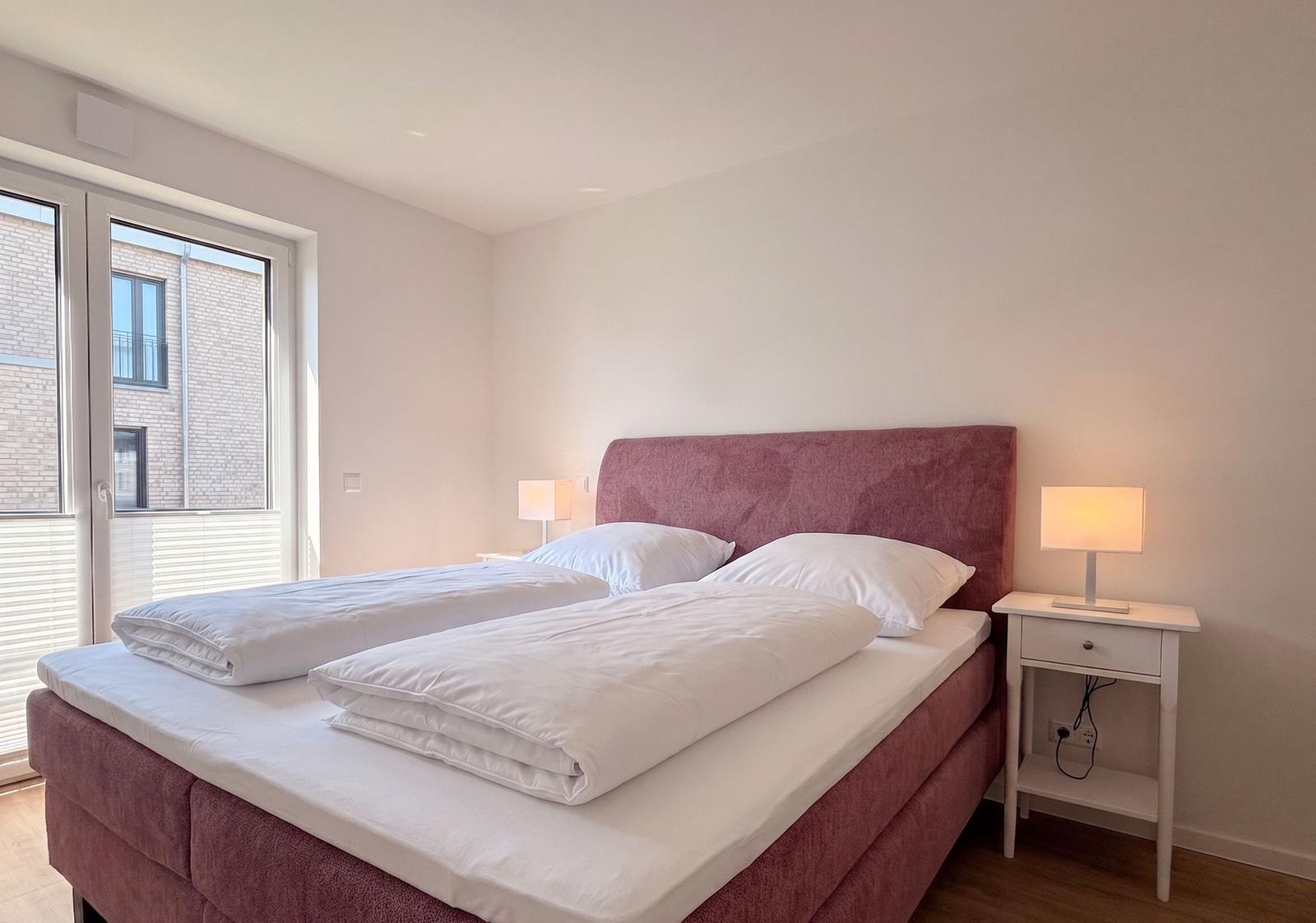 Schlafzimmer mit Bett, Nachttisch und Fenster. Bett mit weißen Bettwäsche.