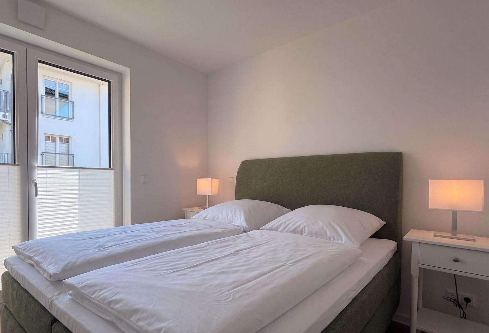 Schlafzimmer mit Bett, Nachttischen und Lampe. Fenster mit Blick auf Gebäude.
