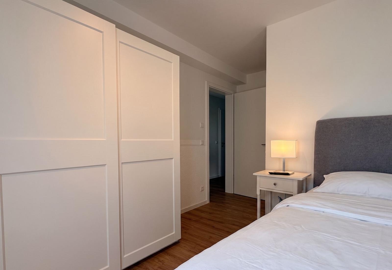 Schlafzimmer mit Bett, Nachttisch und Schrank. Holzboden und Wandbeleuchtung.