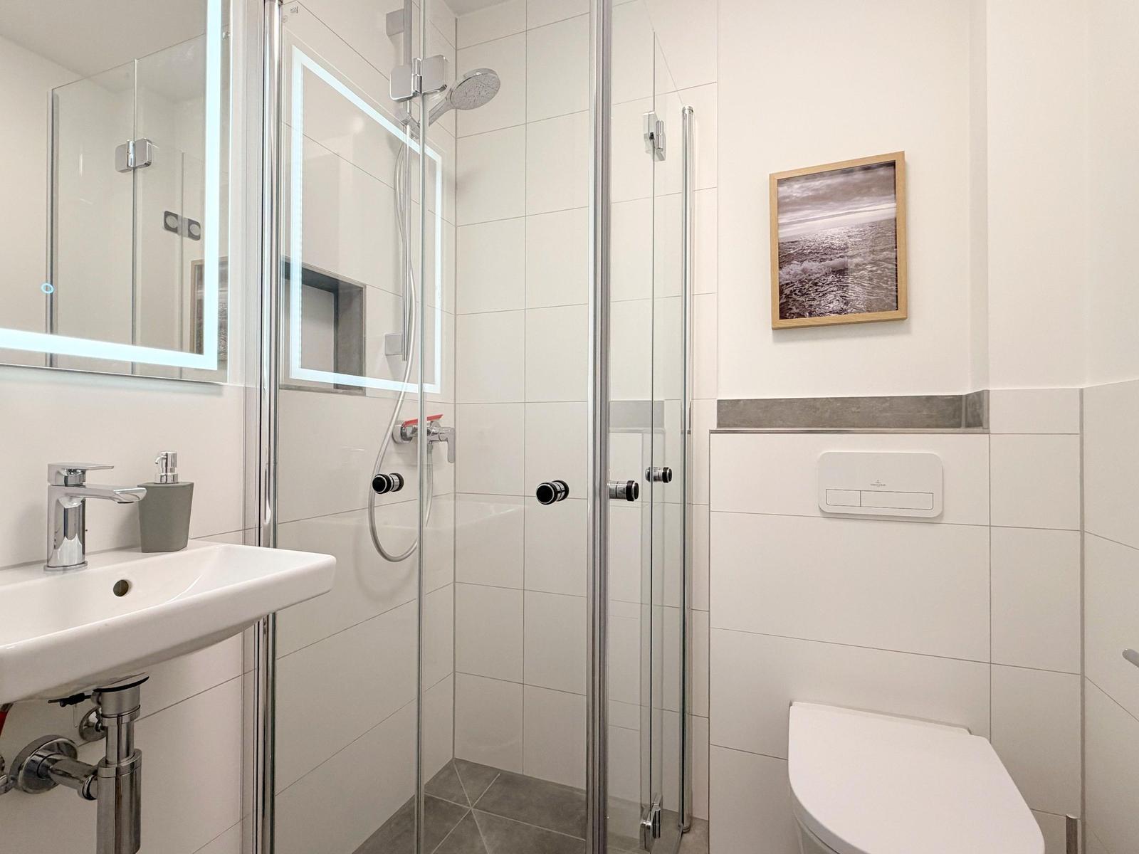 Badezimmer mit Dusche, Waschbecken und Toilette. Wandbild und beleuchtetes Spiegel.