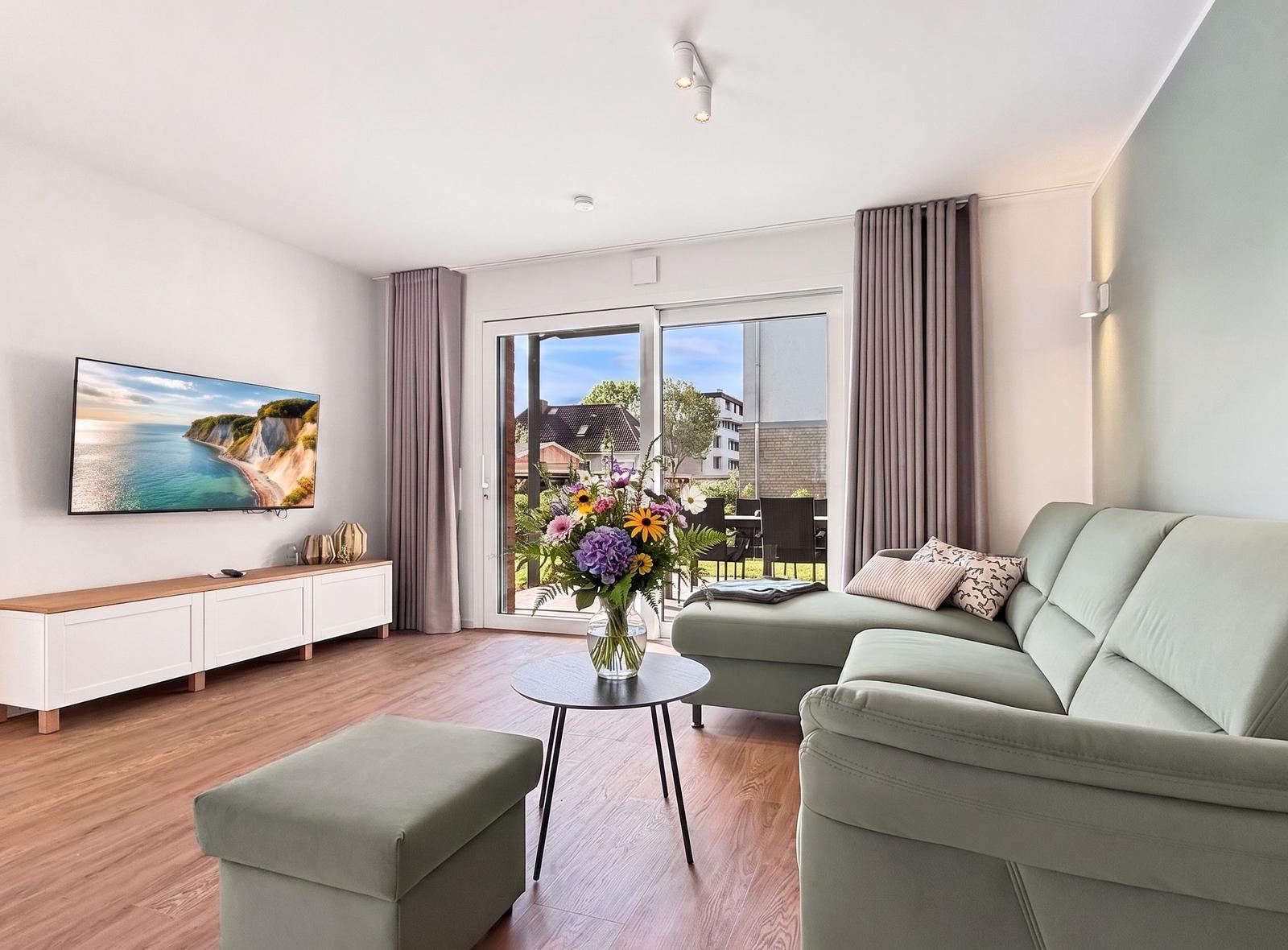 Wohnzimmer mit Sofa, TV und Balkon. Holzboden und Fenster mit Vorhängen.