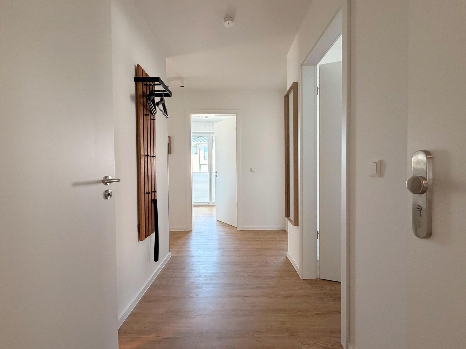 Flur mit Holzboden, weißen Wänden und Türen. An der Wand hängt ein Holzgarderobenständer.