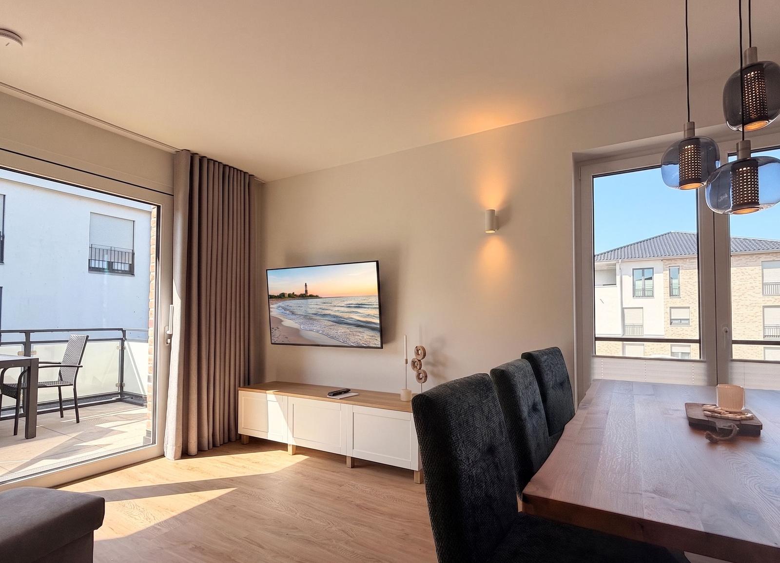 Wohn- und Essbereich mit TV, Esstisch und Balkontür. Blick auf Gebäude und Himmel.