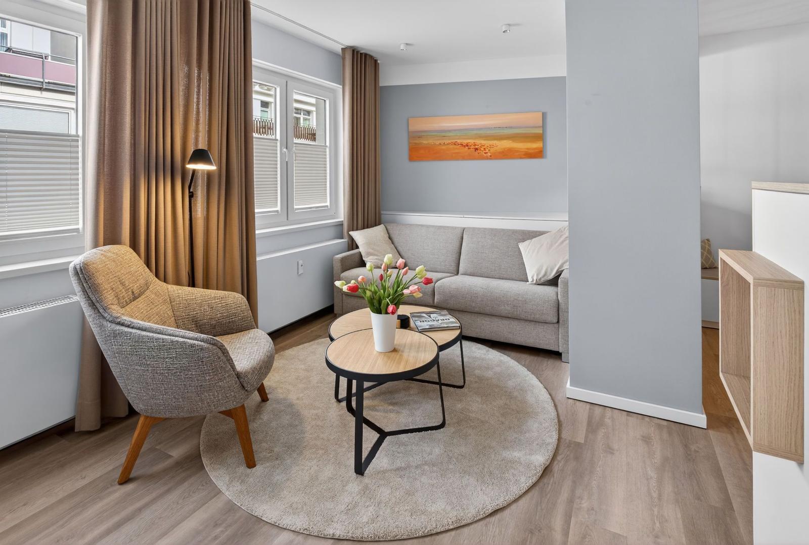 Wohnzimmer mit Sofa, Sessel, Rundteppich und Tisch. Wandbild und Fenster mit Vorhängen.