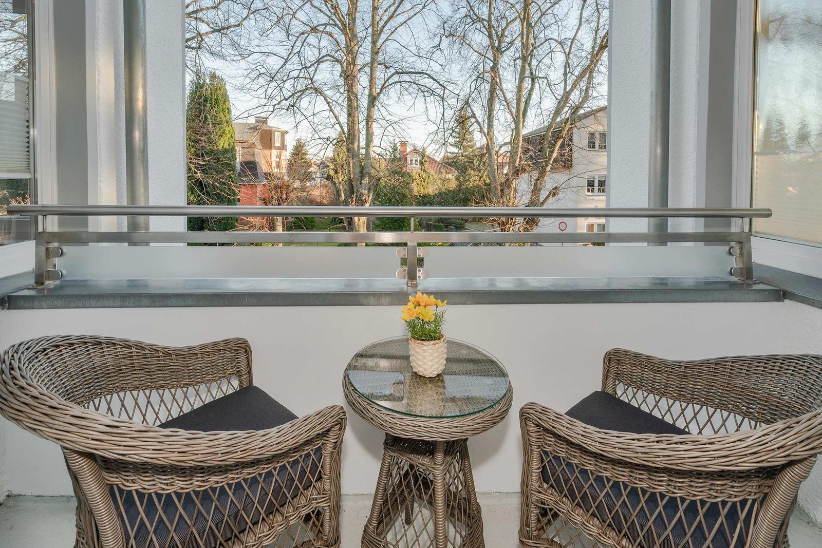 Balkon mit zwei Stühlen und Tisch. Draußen sind Bäume und Häuser sichtbar.