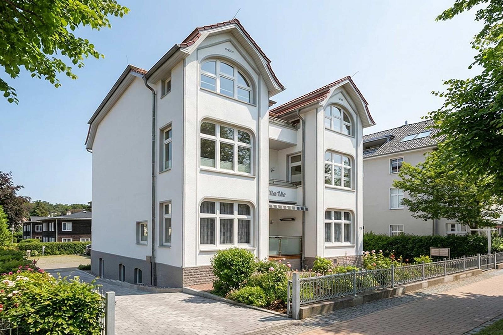 Haus mit weißen Wänden, rotem Dach und Balkon. Umgeben von Grün und Ziergarten.