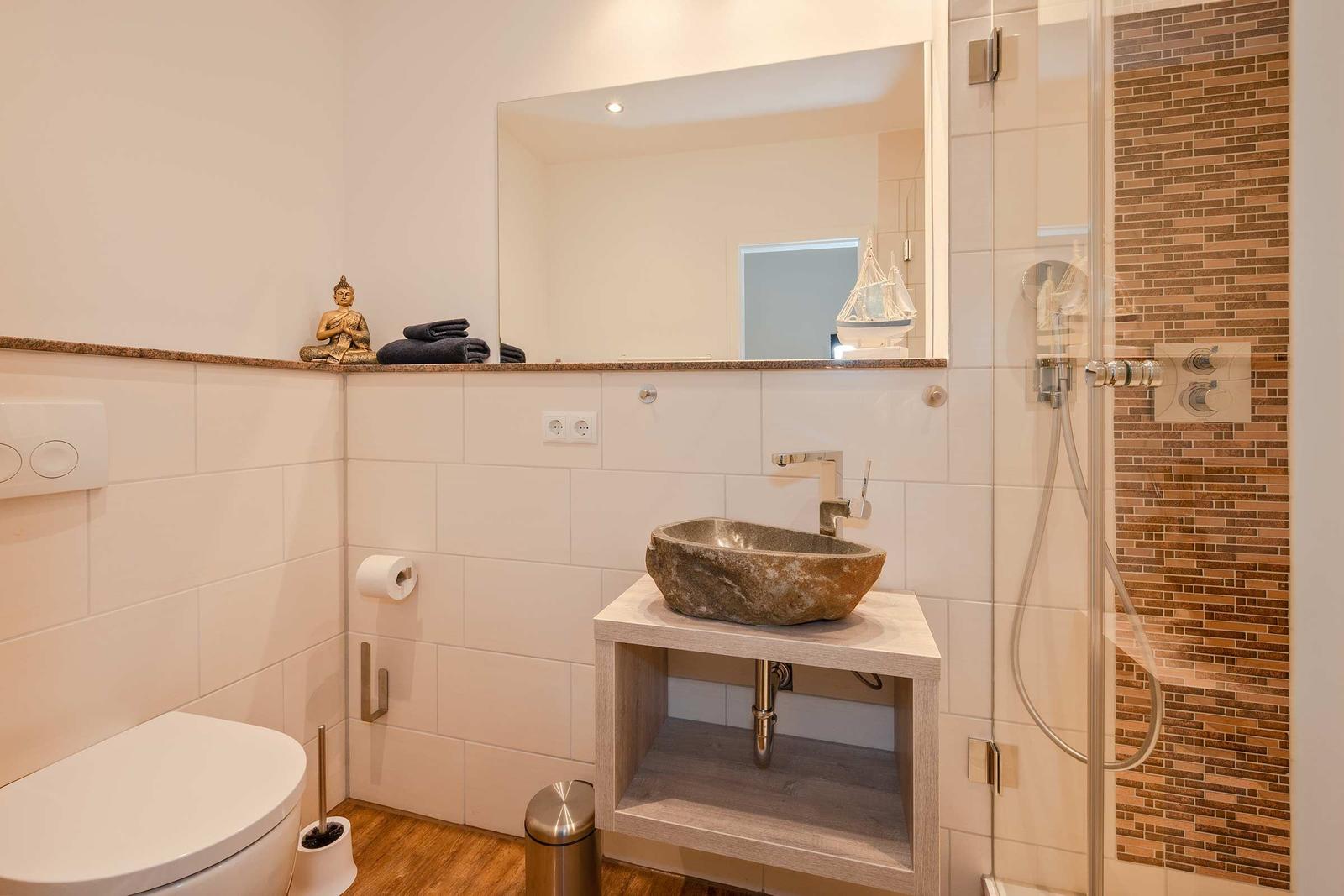 Badezimmer mit Steinwaschbecken, Dusche und Spiegel. Auf der Ablage stehen Handtücher und eine Buddha-Figur.