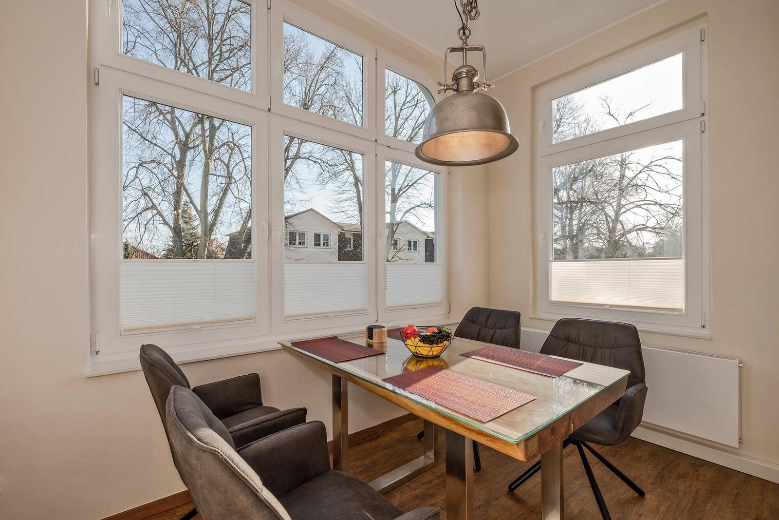 Esszimmer mit Glas-Tisch, dunklen Stühlen und großen Fenstern. Hängelampe und Blick auf Bäume.