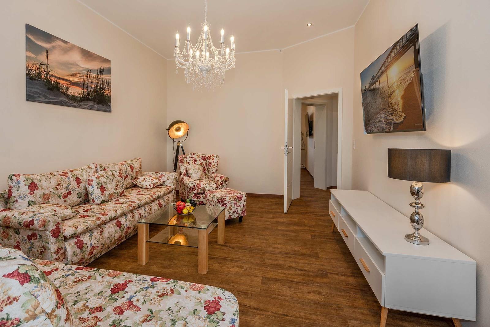 Wohnzimmer mit floralen Sofas, Glas-Tisch und Kronleuchter. Fernseher und Stehlampe an der Wand.