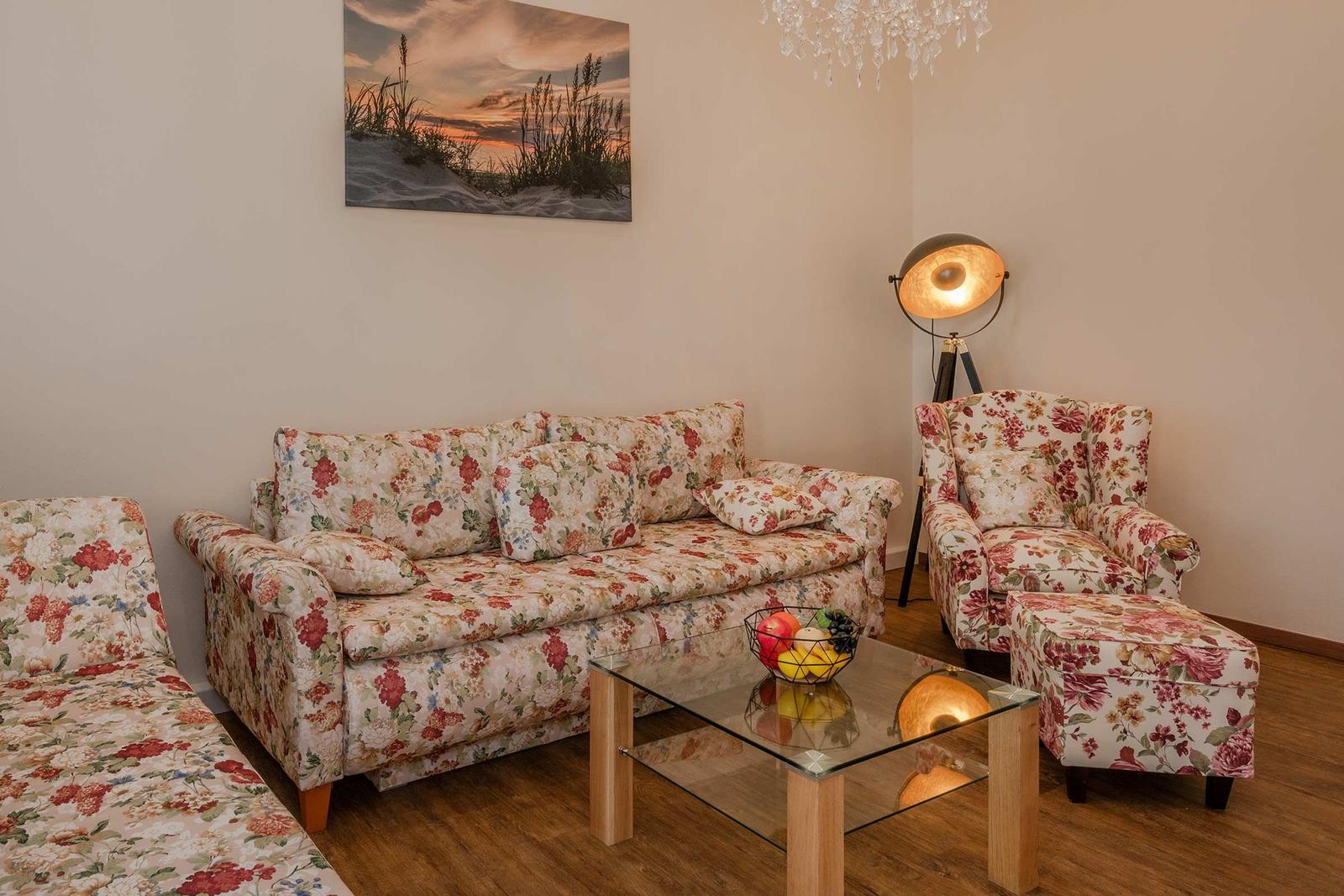 Wohnzimmer mit floralen Sofas, Glas-Tisch und Wandbild.