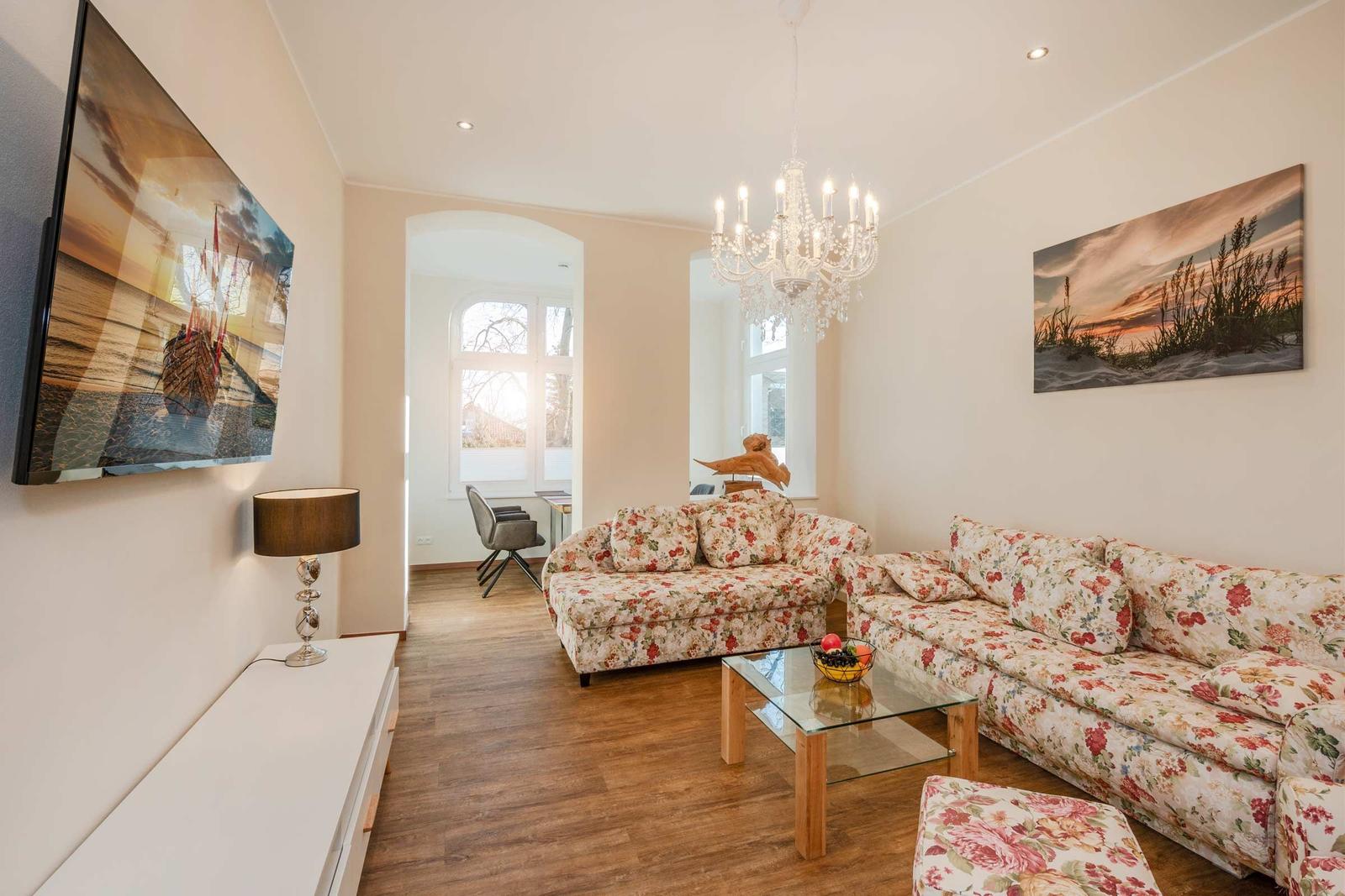 Wohnzimmer mit floralen Sofas, Glas-Tisch und Kronleuchter. Wandbild und Fernseher an der Wand.