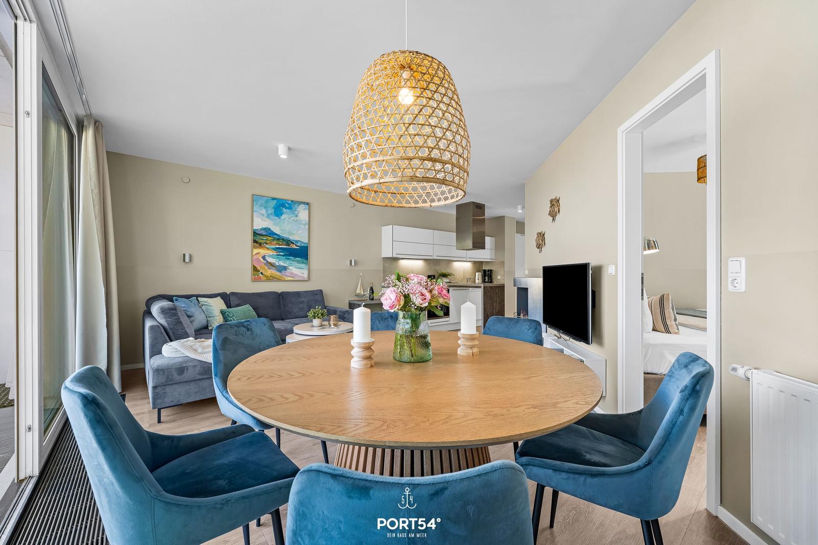 Rundes Esszimmer mit blauen Stühlen und Holztisch. Wohnbereich mit Sofa und Küchenbereich im Hintergrund.