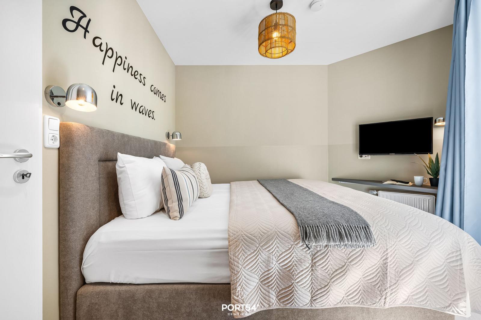 Schlafzimmer mit Bett, Wandbeleuchtung und TV. Wand mit Aufschrift Happiness comes in waves.