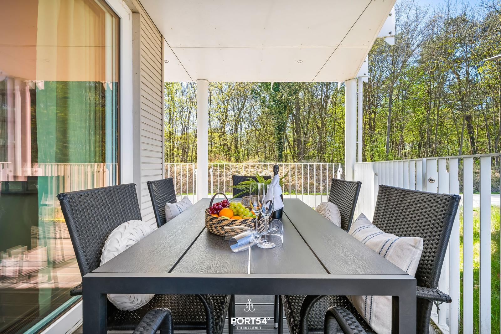Terrasse mit Tisch und Stühlen, Obstkorb und Flasche. Blick in den Wald.