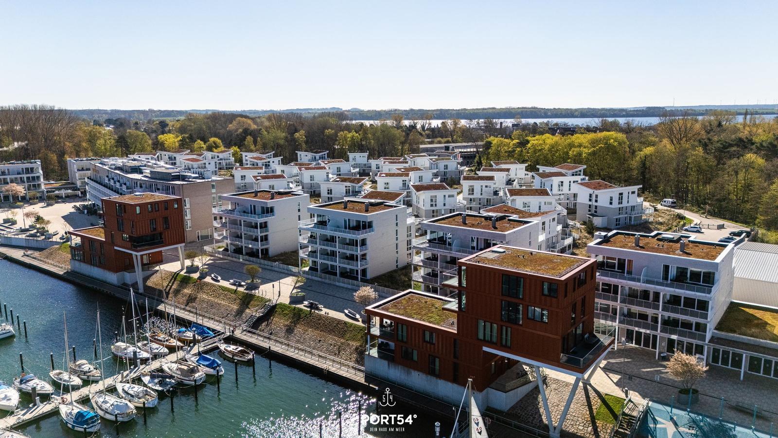 Moderne Wohnhäuser am Hafen mit Booten und grünen Bäumen im Hintergrund.