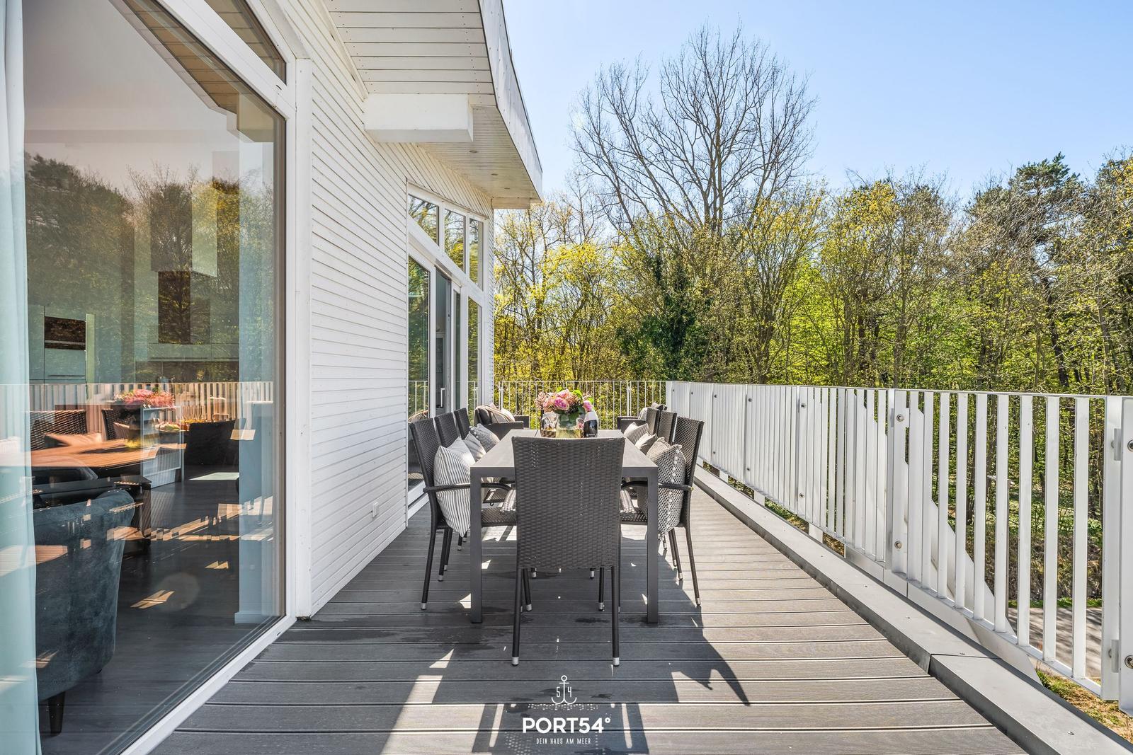 Terrasse mit Tisch und Stühlen, Blick in den Wald. Glasfront und weißer Geländer.