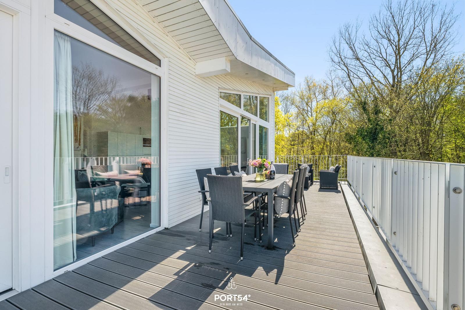 Terrasse mit Tisch und Stühlen, Blick auf Bäume und Haus mit Glasfront.