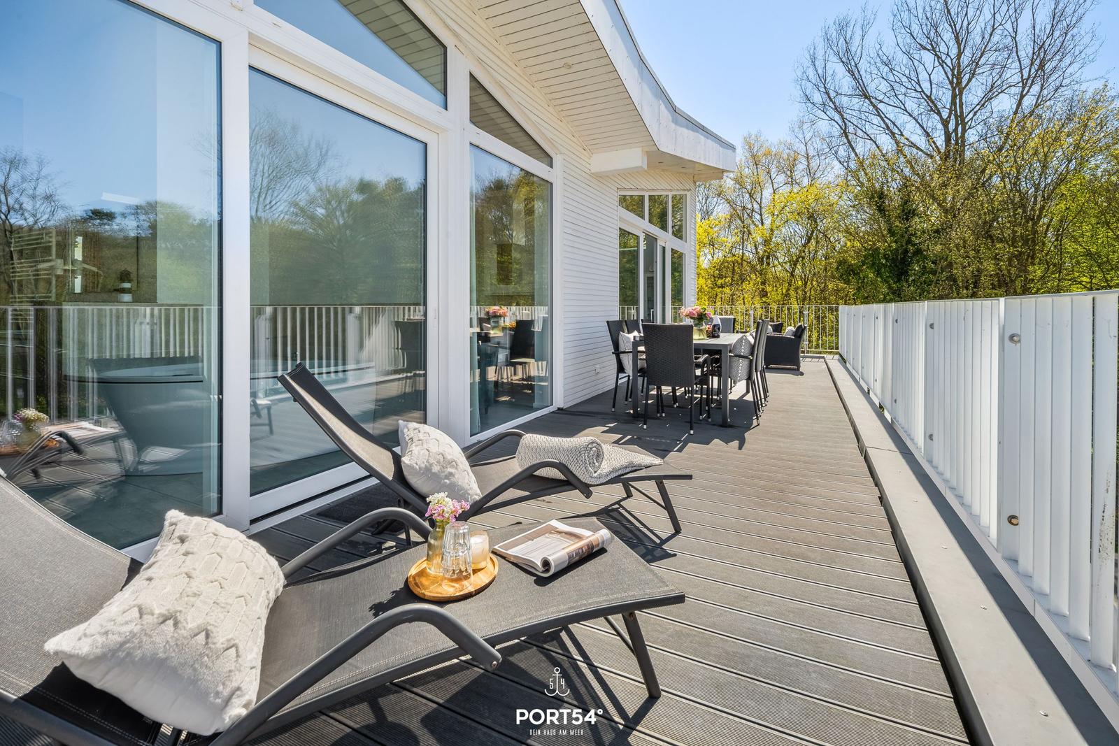 Terrasse mit Liegestühlen, Tisch und Stühlen. Weiße Hauswand und Glasfront. Hintergrund mit Bäumen.