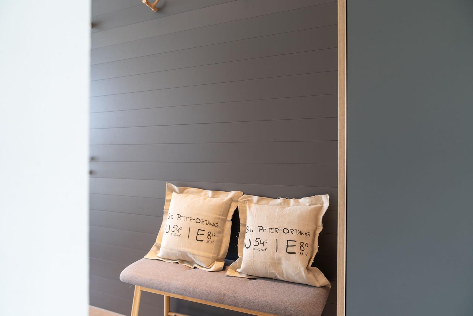 Zwei Kissen mit Ortsangaben liegen auf einer Bank vor einer Holzwand.