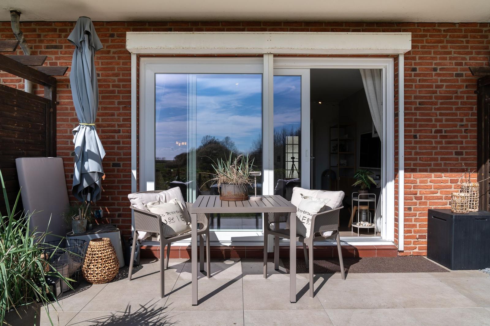 Terrasse mit Tisch, zwei Stühlen und Sonnenschutz. Durchgang ins Haus mit Blick auf Garten.