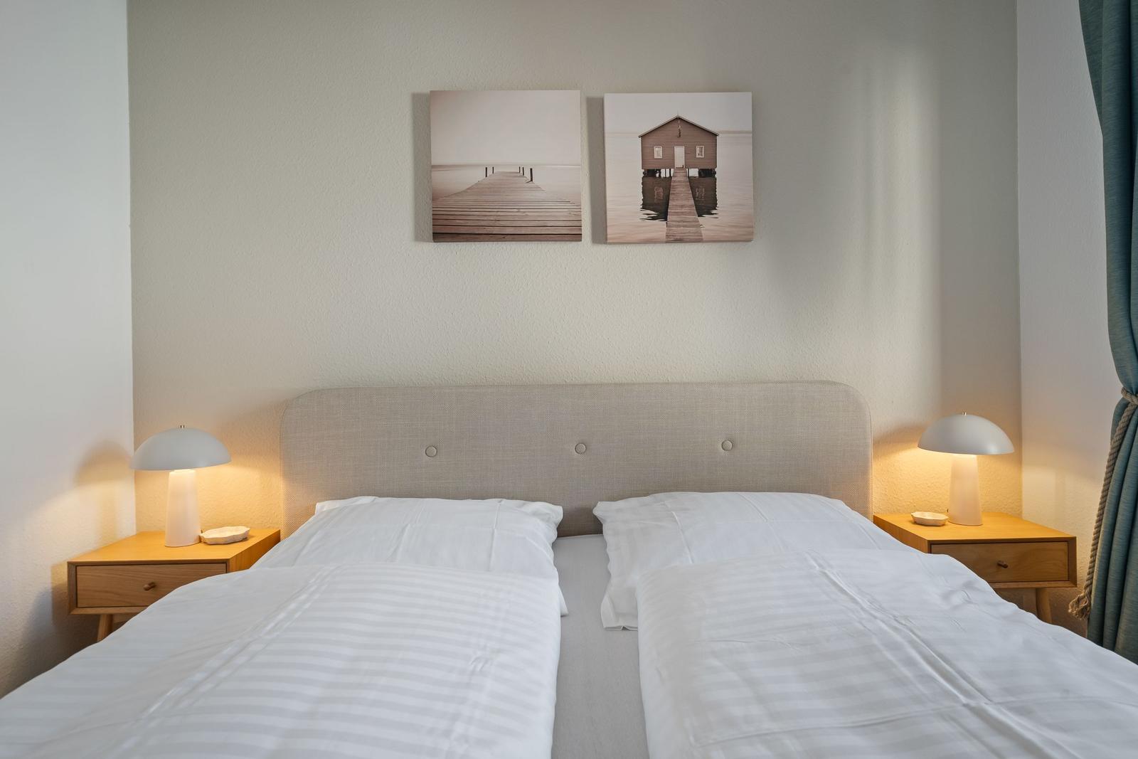 Doppelbett mit weißen Bettwäsche, zwei Nachttische und Wandbildern.