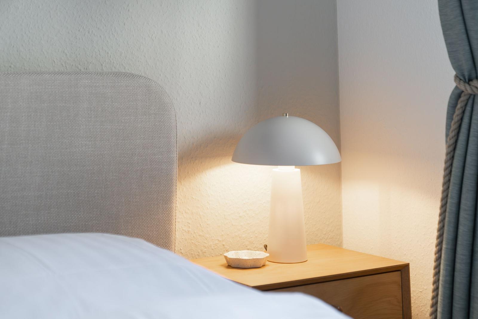 Schlafzimmer mit Nachttisch, Lampe und Bett. Wand und Vorhang sind sichtbar.