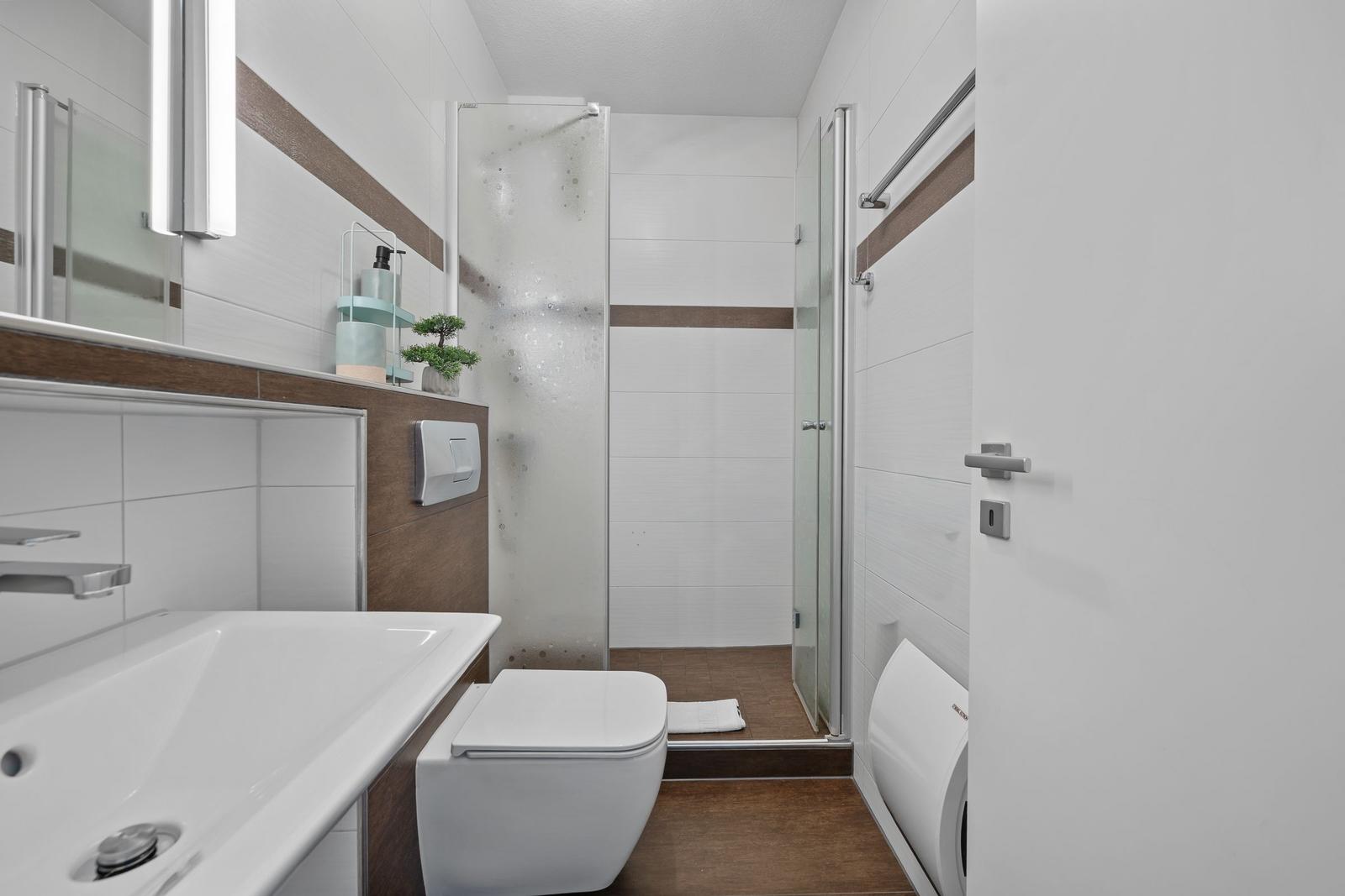 Bad mit Dusche, Waschbecken und Toilette. Holzakzent an Wand und Boden.
